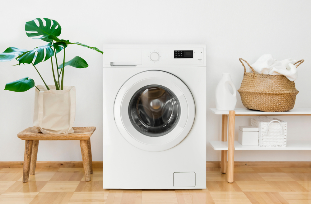 A white washing machine sitting in between a plant and some shelves